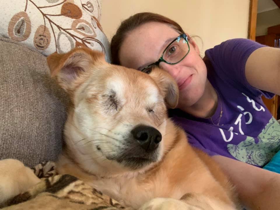 Woman in glasses and a purple shirt resting her head against a sleeping tan terrier mix on a couch.